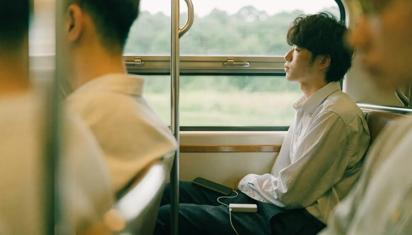 電車の窓際でモバイルバッテリーからスマホを充電しながら景色を眺める男性のイメージ